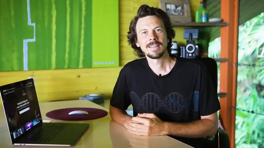 A man seated at a desk in a brightly lit room with green and yellow walls, speaking while gesturing with his hands. A laptop displaying a website is open on the desk, and a camera is visible on a shelf behind him. Natural light streams in from a window on the right.