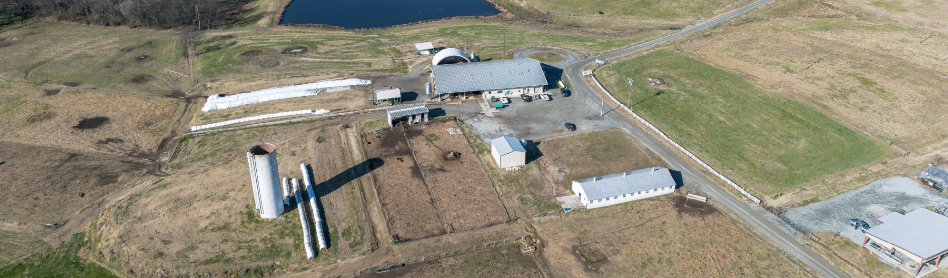 Aerial view of a farm complex featuring several white outbuildings, a tall silver silo with wrapped hay bales nearby, fenced livestock pens, and a large pond in the background under a clear sky.