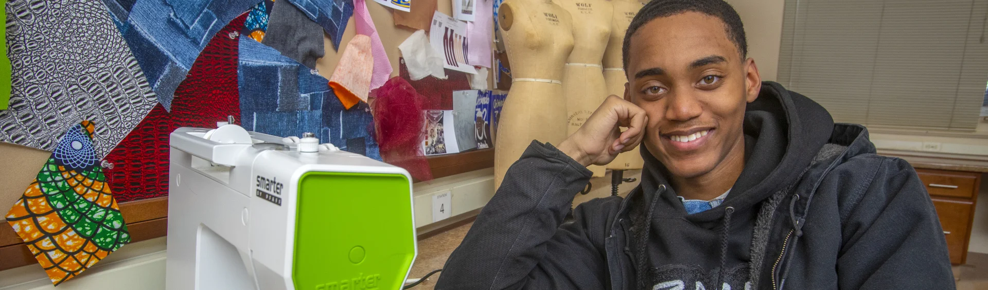 A smiling student sits at a table in a sewing classroom beside a sewing machine. Behind him, a mannequin and a bulletin board display colorful fabric samples and patterns.