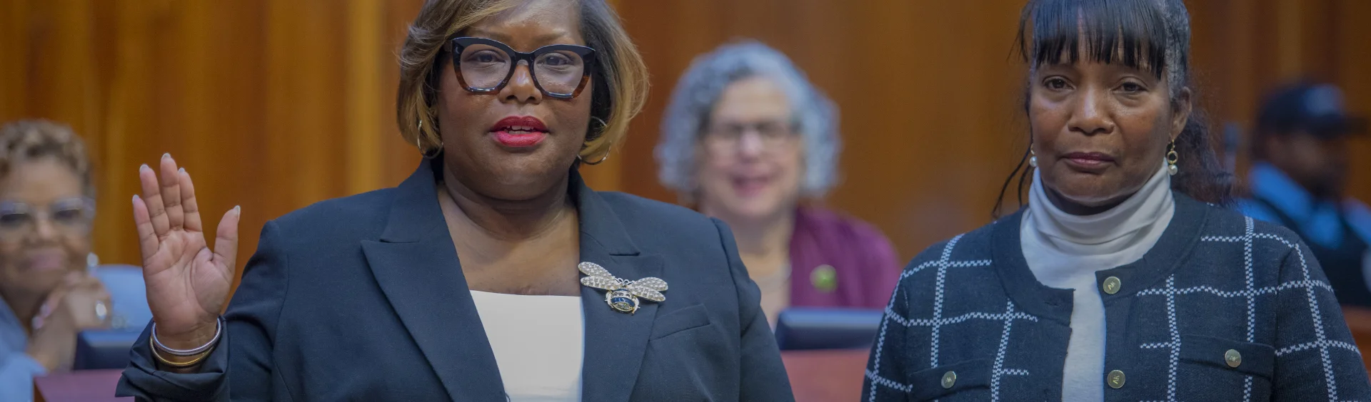 A woman raises her right hand while taking an oath beside another woman during a formal ceremony. Both stand in front of seated attendees in a wood-paneled room.