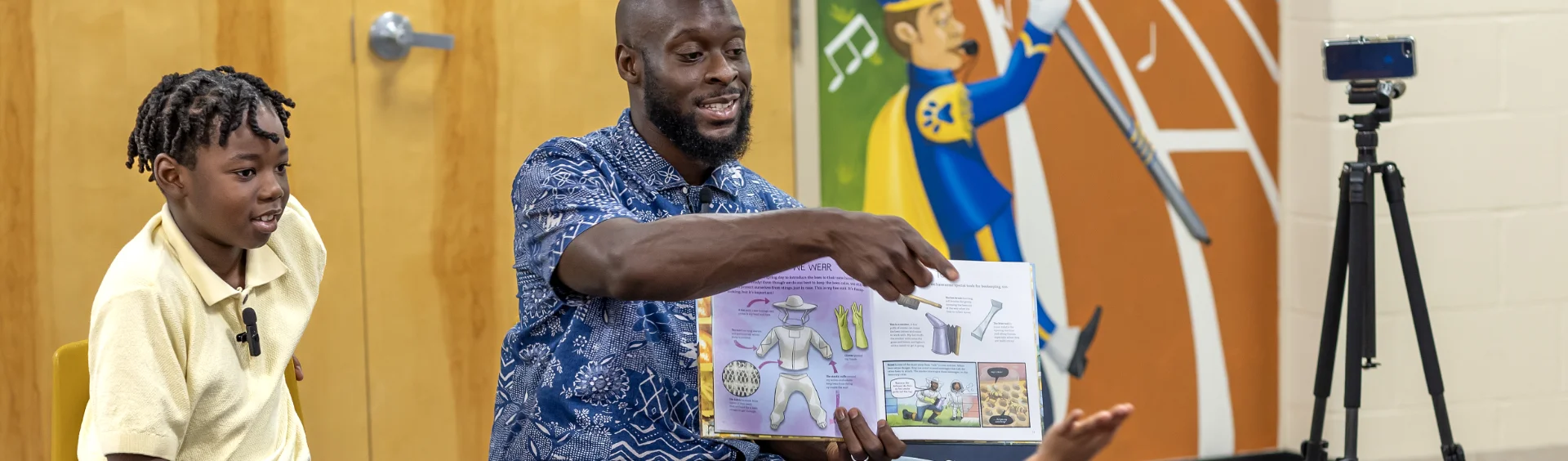 A man sits next to a child while reading aloud from a picture book to an unseen audience. The man gestures toward the open pages showing colorful illustrations. Behind them, a wall mural depicts a marching band musician, and a camera on a tripod records the session.