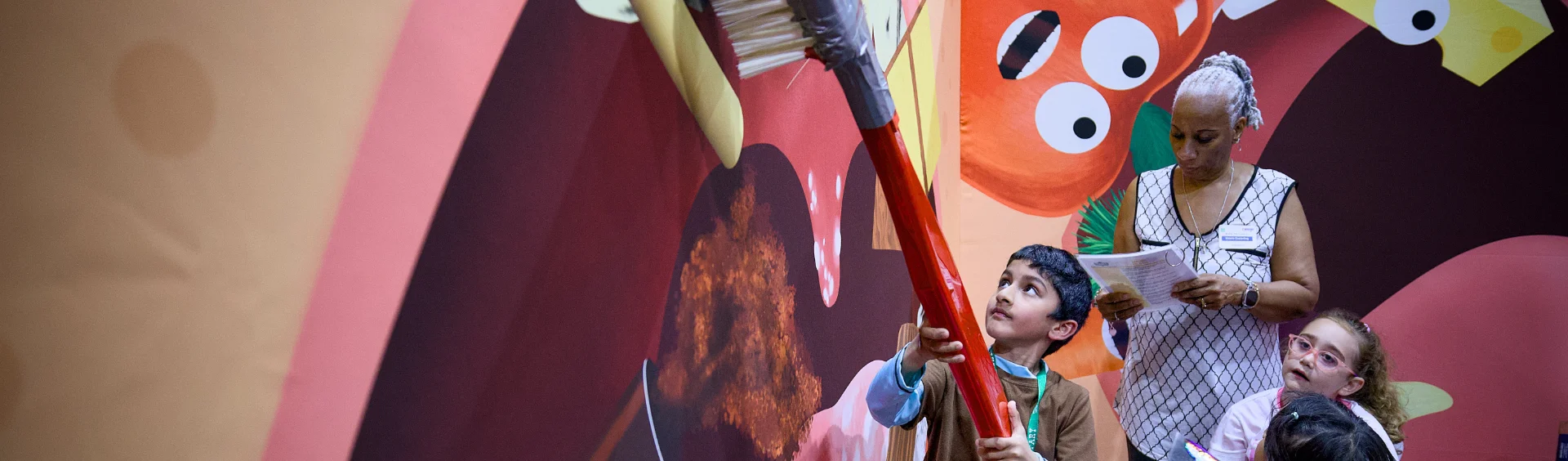 A young boy holds a giant red toothbrush prop in front of a brightly colored backdrop featuring cartoon food characters. A woman standing behind him looks down at some papers, while another young child with glasses looks on from the bottom right.
