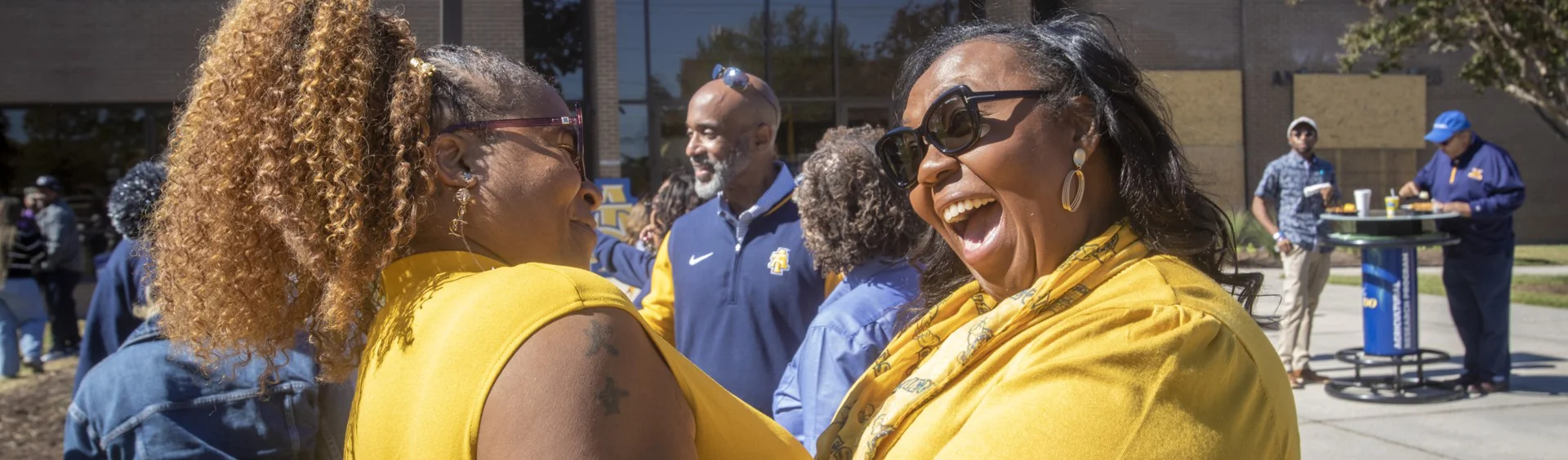 Two women wearing yellow tops smile and laugh together outdoors at a social event,with people talking and eating in the background near a table.