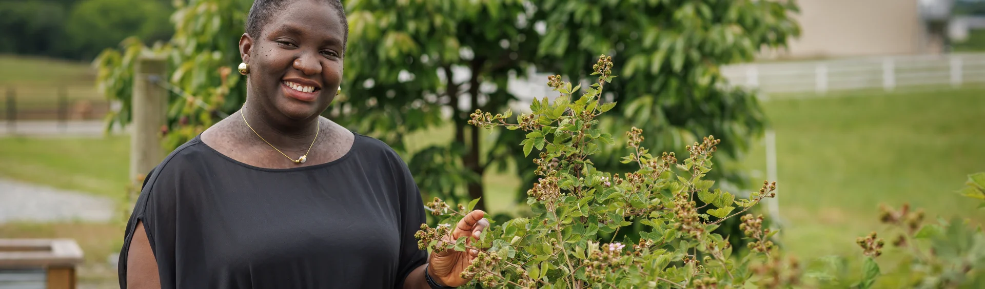 A smiling Black woman in a black shirt stands outdoors, gently holding a branch of a berry bush with a farm landscape in the background.