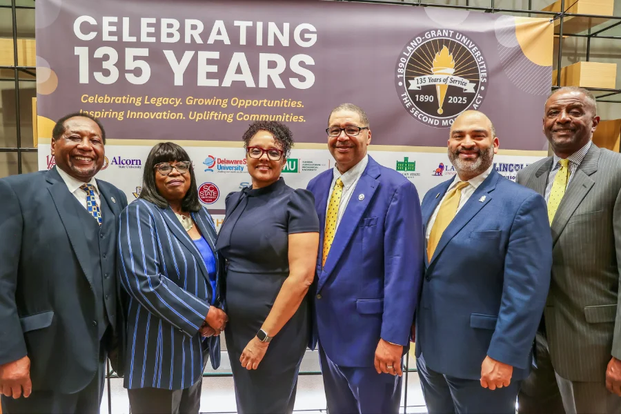 Six people stand together smiling in front of a large banner that reads “Celebrating 135 Years” with the 1890 Land-Grant Universities logo. The banner includes the text “Celebrating Legacy. Growing Opportunities. Inspiring Innovation. Uplifting Communities.” The group is dressed in professional attire and standing indoors at an event celebrating the Second Morrill Act anniversary.