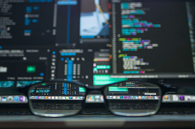 Close-up of eyeglasses resting on a desk, with computer monitors displaying blurred lines of colorful code in the background